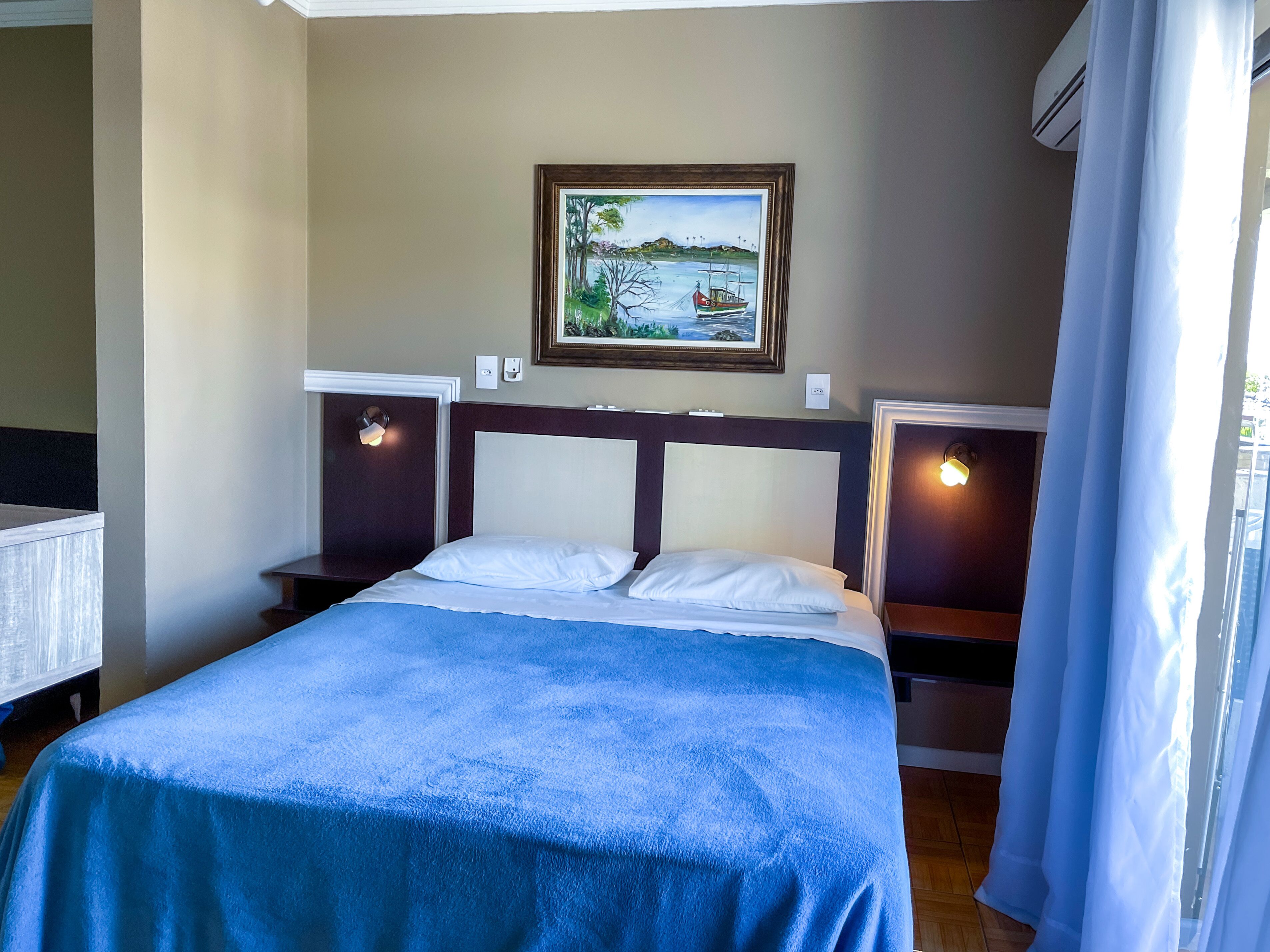 Une chambre d’hôtel comprenant un lit, deux tables de chevet, une commode, un tableau au mur et un appareil de climatisation.