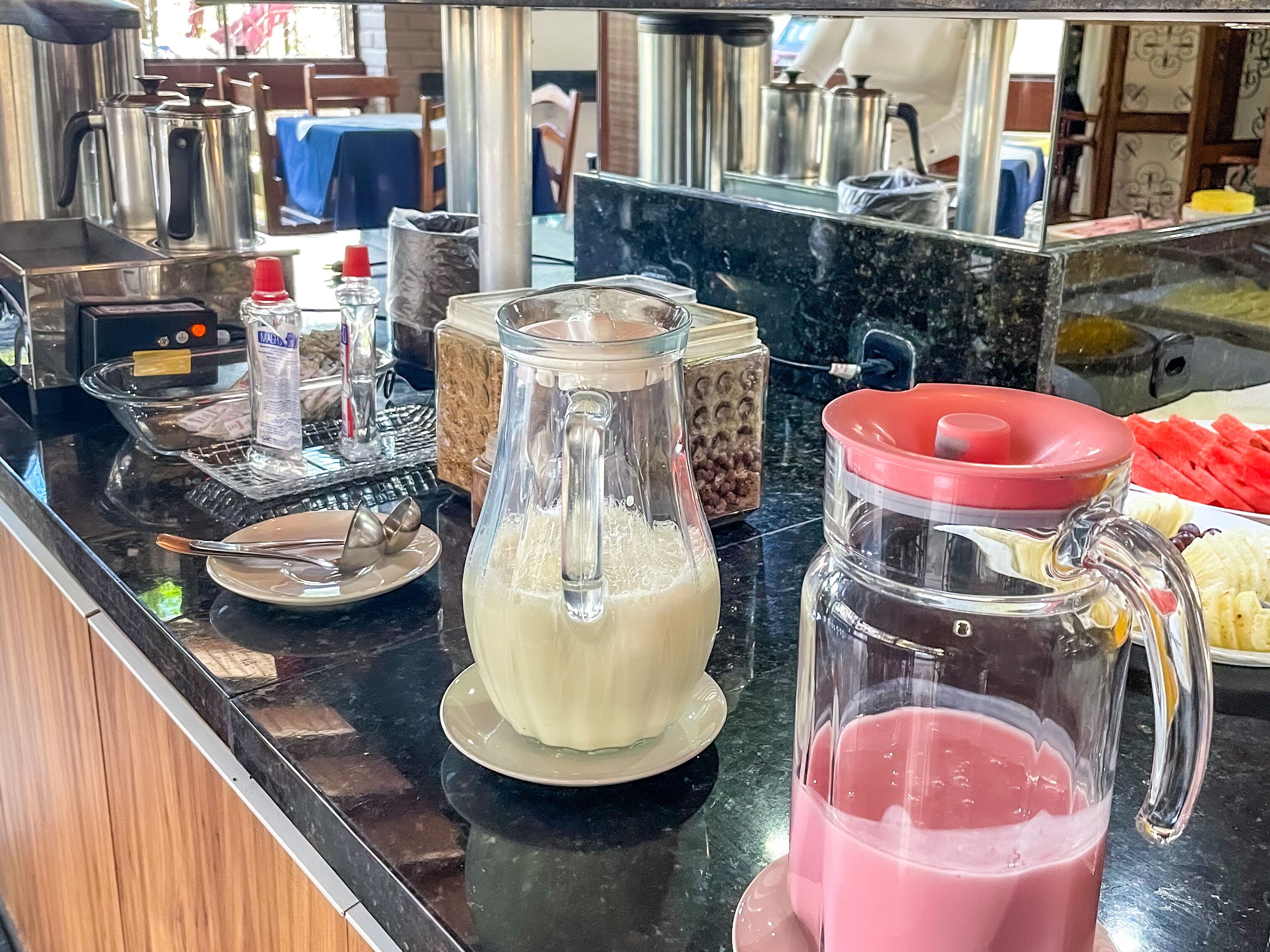 Una encimera de cocina con una cafetera, un espumador de leche, una jarra de leche y una licuadora con una mezcla rosada, además de un plato de plátanos en rodajas.