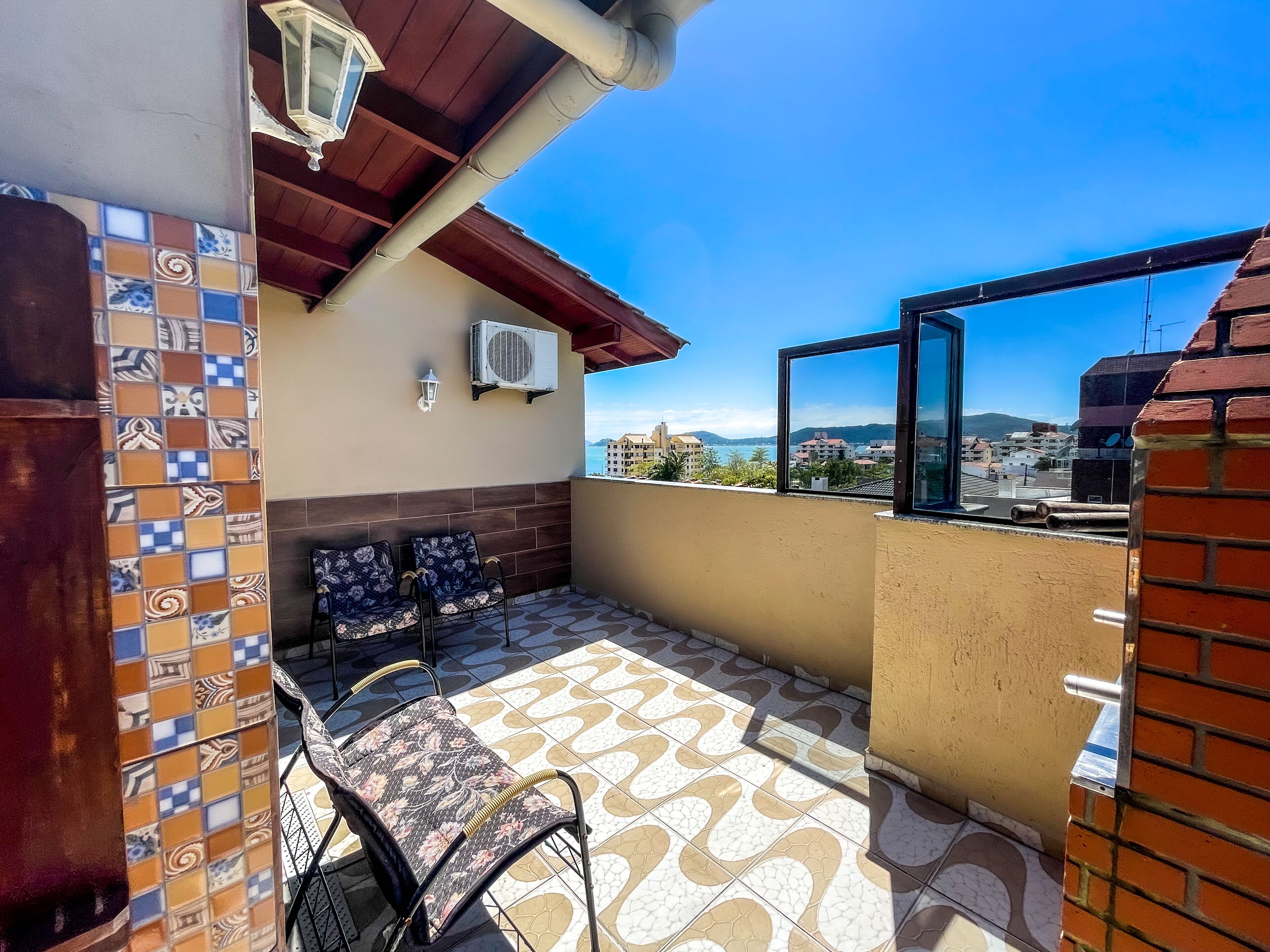 Un balcon orné de carreaux décoratifs, deux fauteuils en osier, une table, un appareil de climatisation et une vue sur des immeubles et des montagnes.