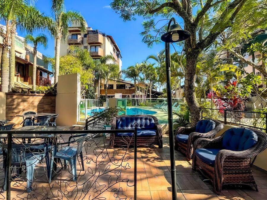 Zona junto a la piscina con mobiliario de mimbre, una mesa y sillas, rodeada de palmeras y vistas a una piscina y edificios.