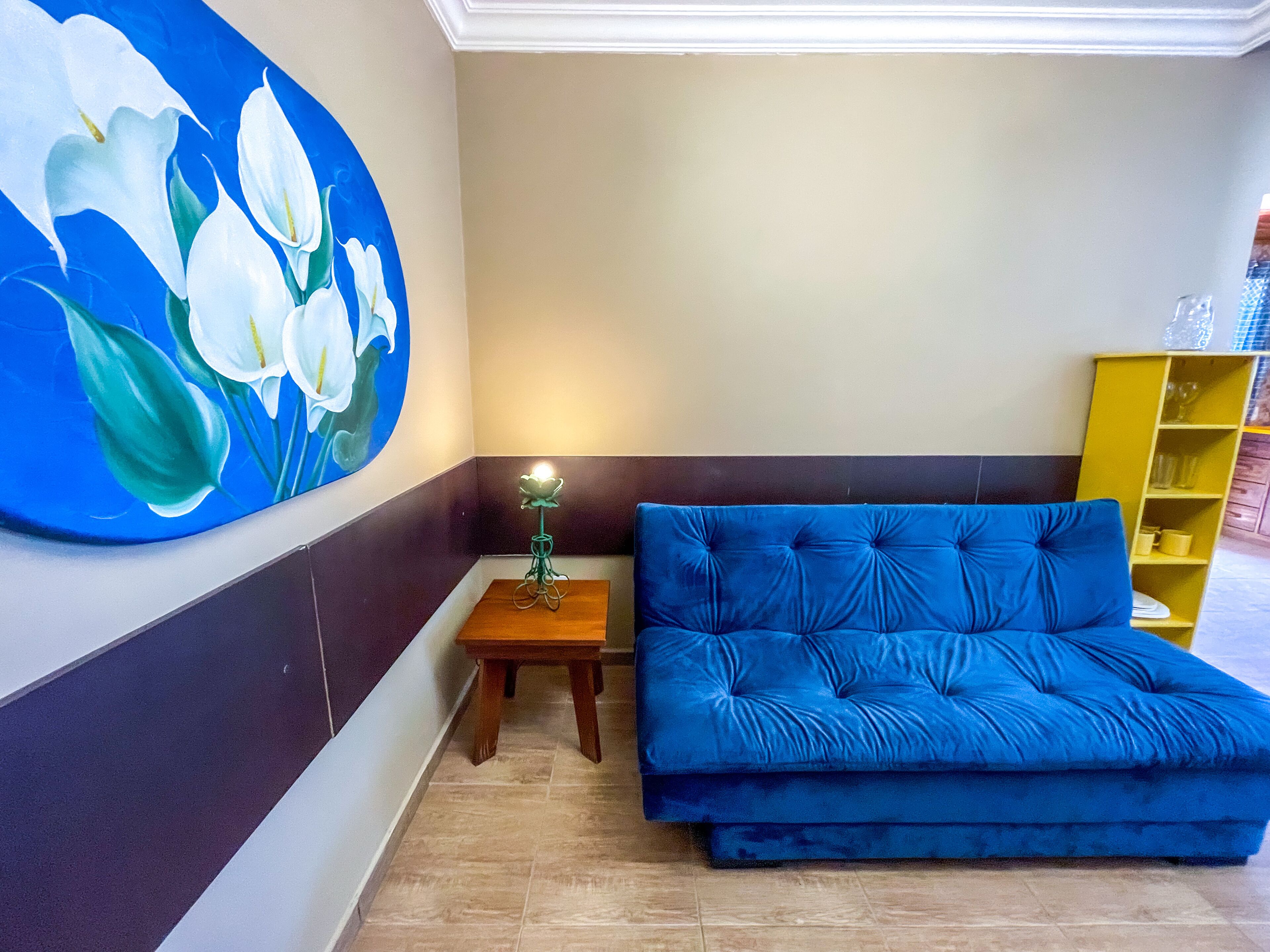 Una stanza con un divano blu, un tavolino in legno con una lampada, una scaffalatura gialla e un grande quadro floreale appeso al muro.