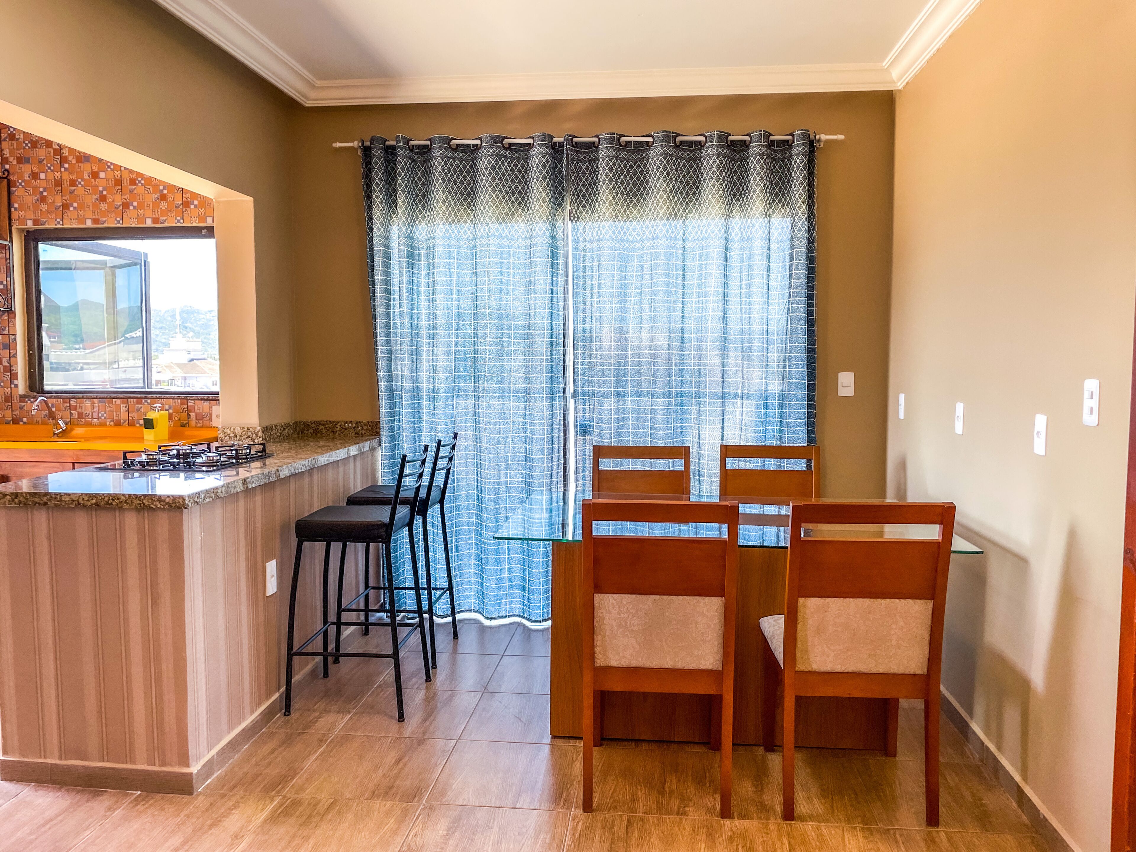 Una cocina con estufa a gas, un lavamanos y una ventana con cortinas estampadas. Se observan dos sillas de madera y una mesa pequeña.