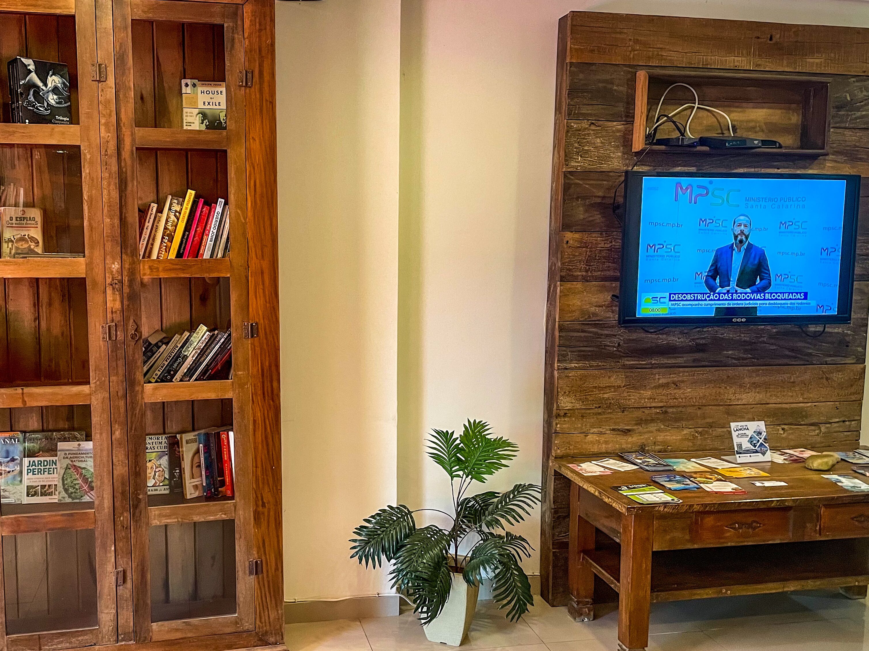 A room with a wooden bookshelf filled with books, a television mounted on a wooden wall displaying a news channel, and a small wooden table with magazines and a plant.