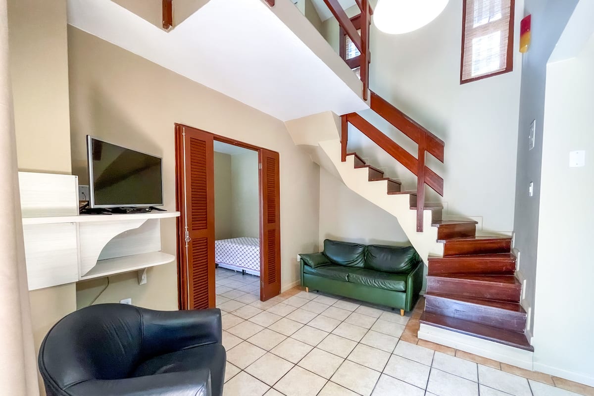 Un salón con una butaca de cuero negro, un televisor en un estante, un sofá verde y una escalera con barandillas de madera.
