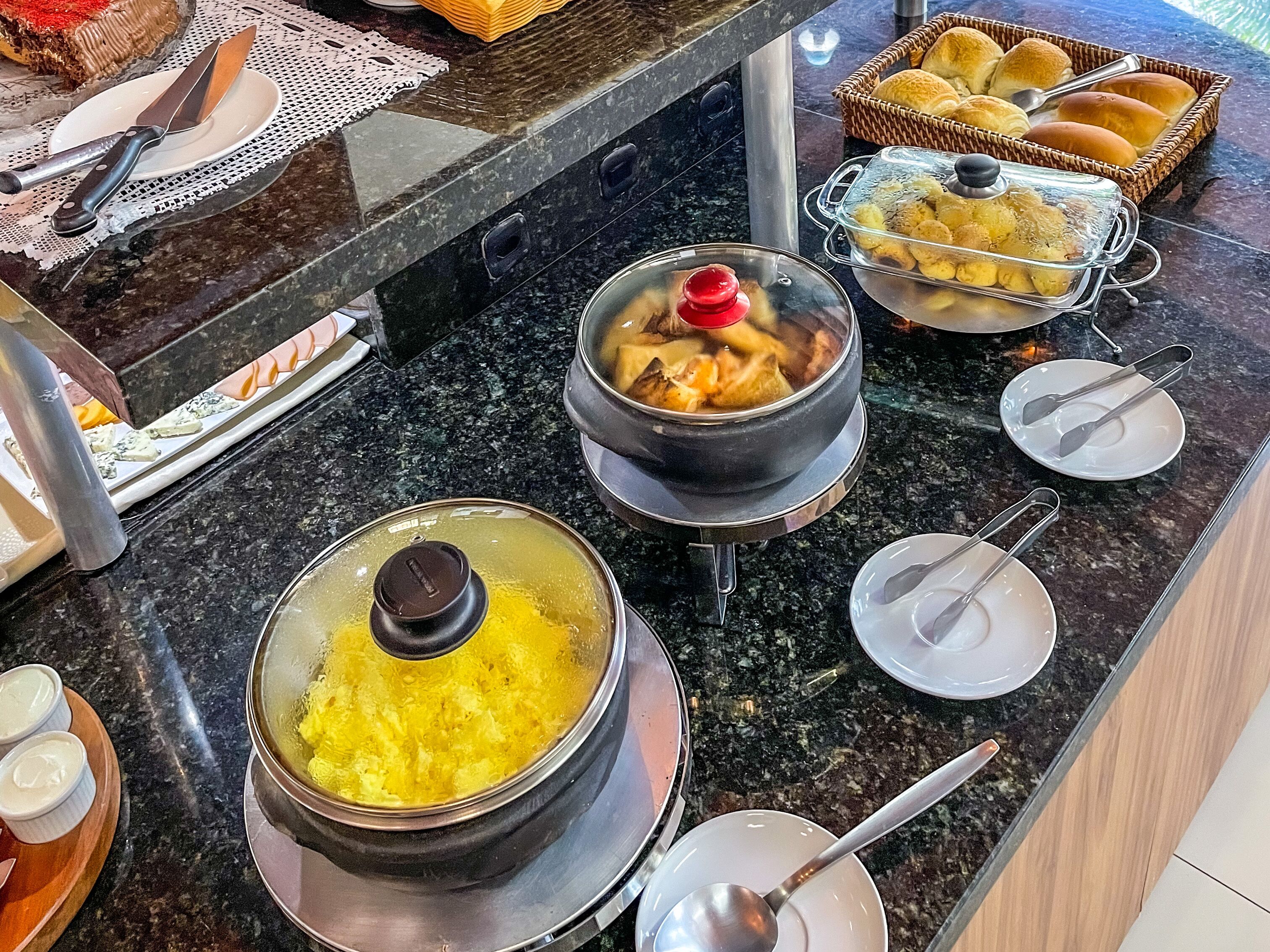 A buffet setup with various dishes, including a pot of soup, a container of yellow food, and a basket of rolls.