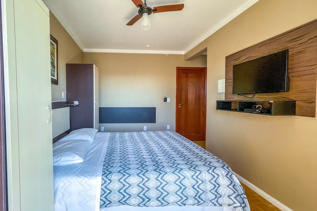 Un dormitorio con cama, ventilador de techo, televisor montado en la pared y una puerta.