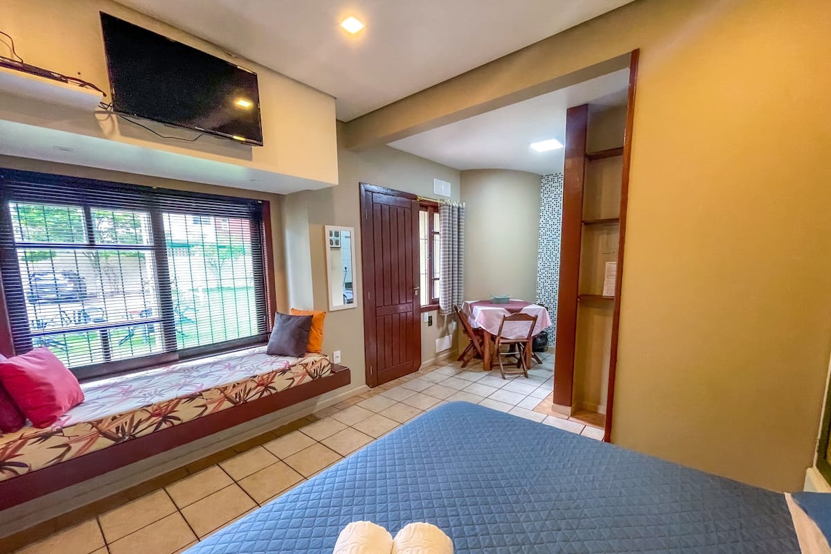 Un dormitorio con cama, una ventana con persianas, una pequeña mesa con sillas y una puerta que da a otra habitación.