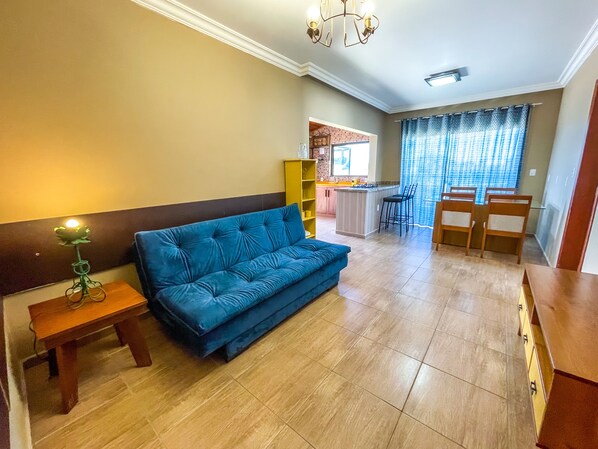 A living room with a blue sofa, a small table with a lamp, and a kitchen area with a bar stool and chairs. - Hotel e Pousada Sonho Meu (Florianópolis)