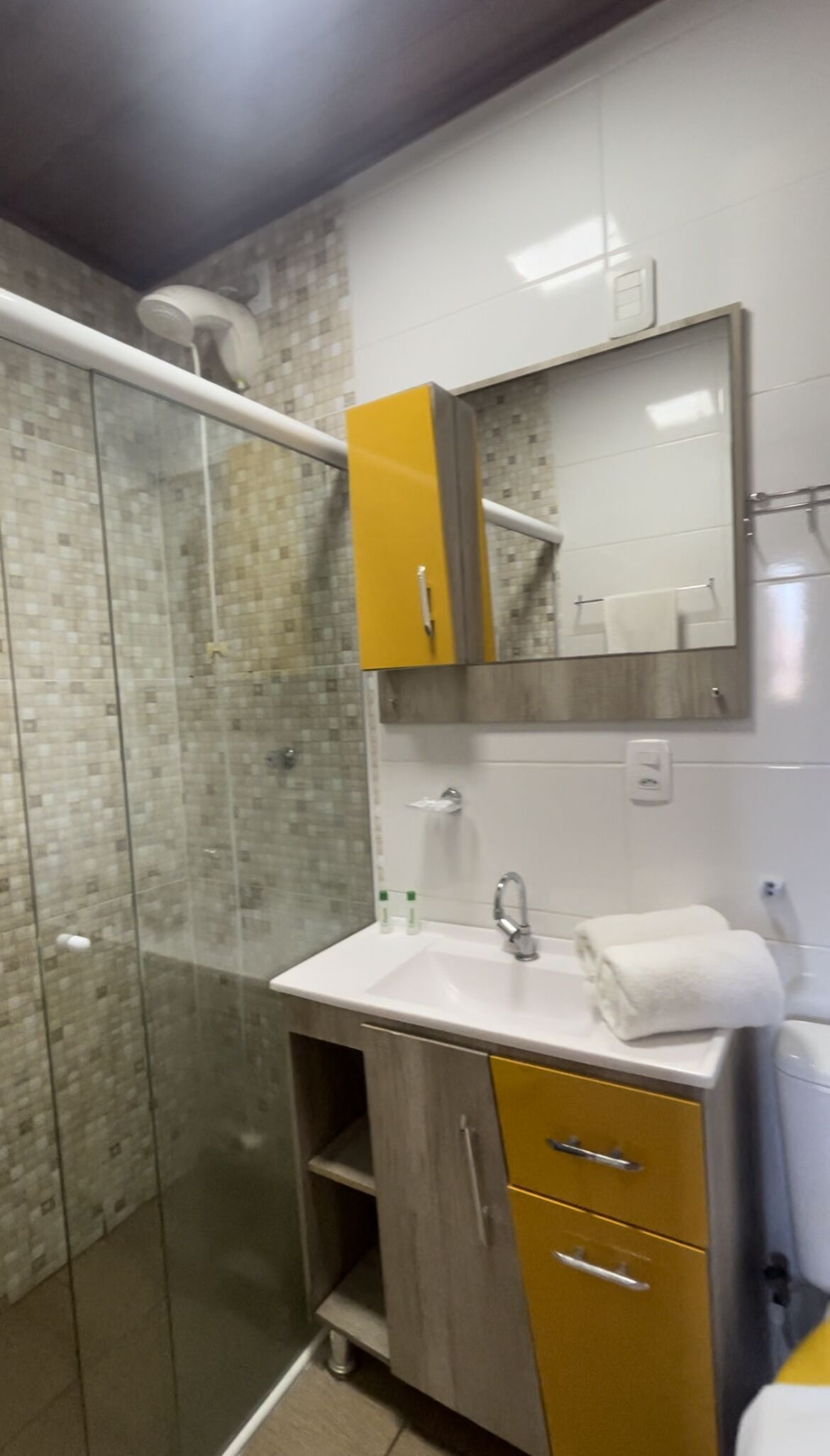 Un baño con cabina de ducha de vidrio, un lavamanos blanco con un espejo encima y un mueble amarillo debajo.