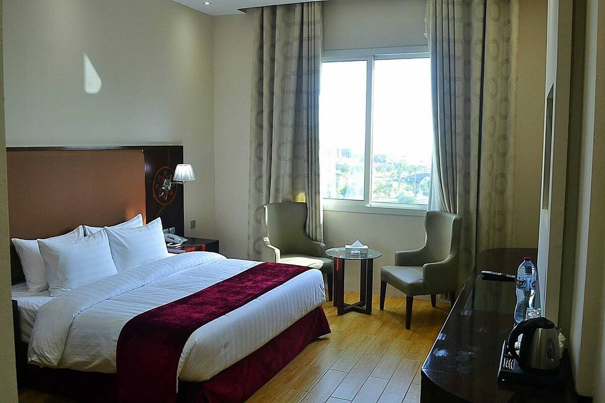 Habitación de hotel con cama, escritorio, dos sillas, ventana con cortinas y vista a la ciudad.