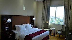 A hotel room with a large bed, two bedside lamps, a small table, a chair, and a view of the city through the window. - Palm Inn Hotel Doha (Doha)