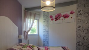 A bedroom with a bed, a window with curtains, a wall with a floral painting, and a patterned wallpaper.