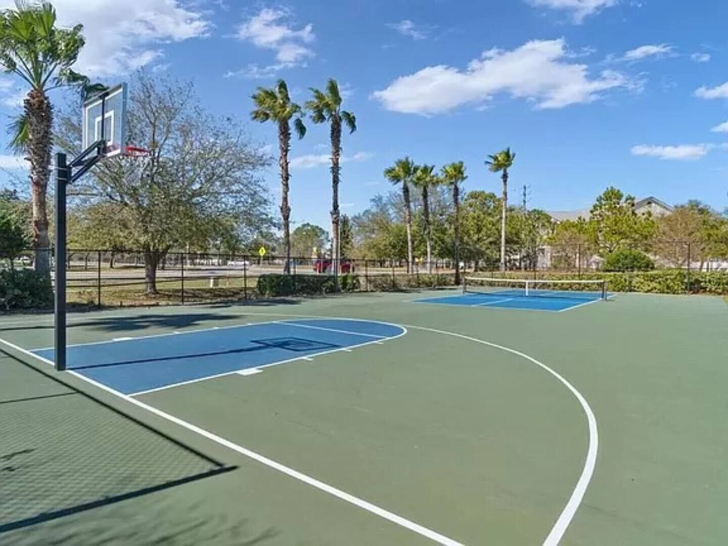An outdoor basketball court with a basketball hoop, surrounded by palm trees and a clear sky.