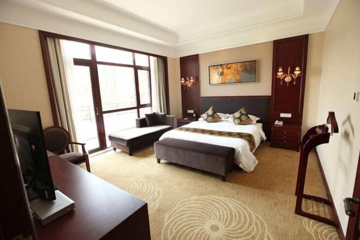 A hotel room with a large bed, a desk with a computer, a chair, a sofa, and a large window.