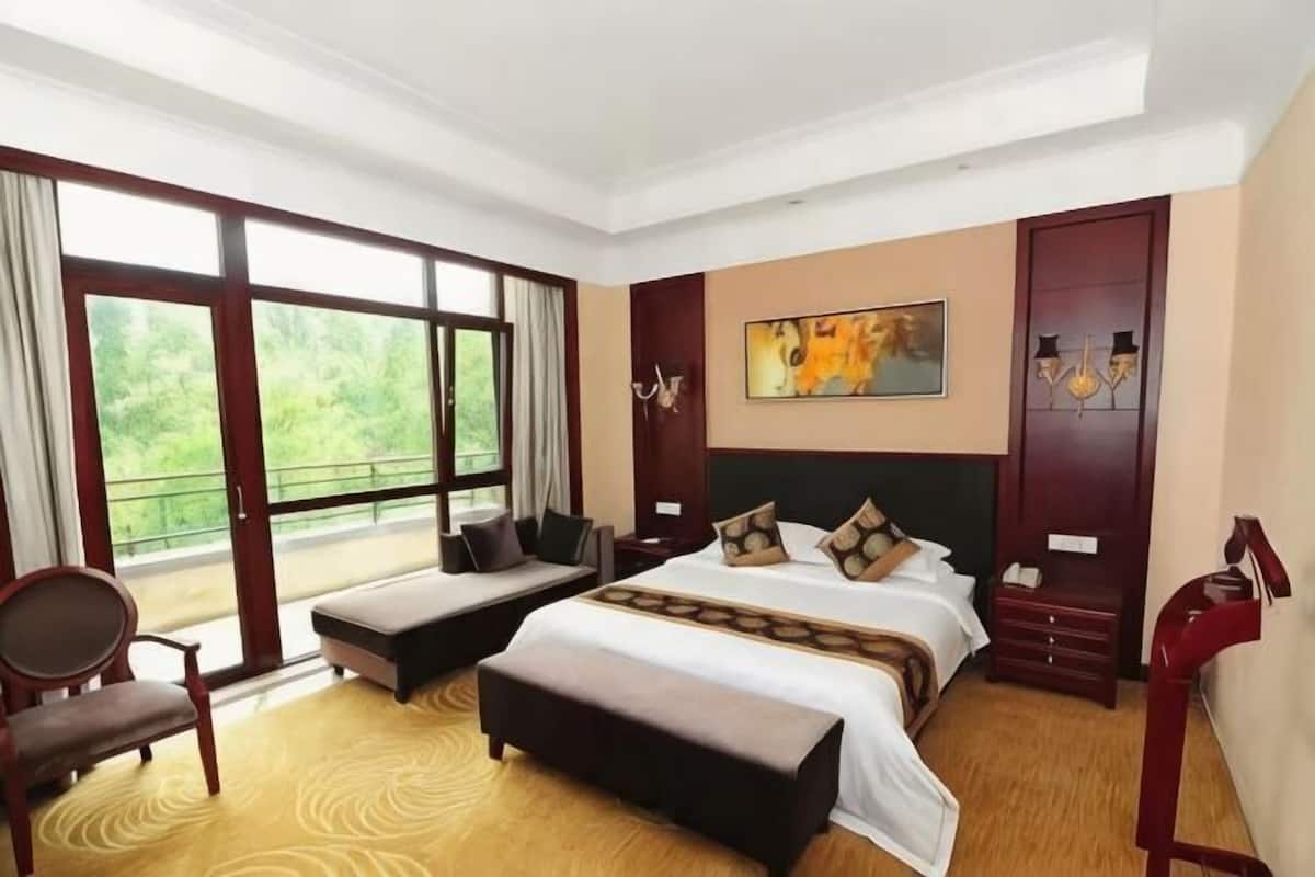 Ein Hotelzimmer mit einem großen Bett, einer Bank, einem Sessel, einem Schreibtisch und Blick auf Grün durch die Fenster.