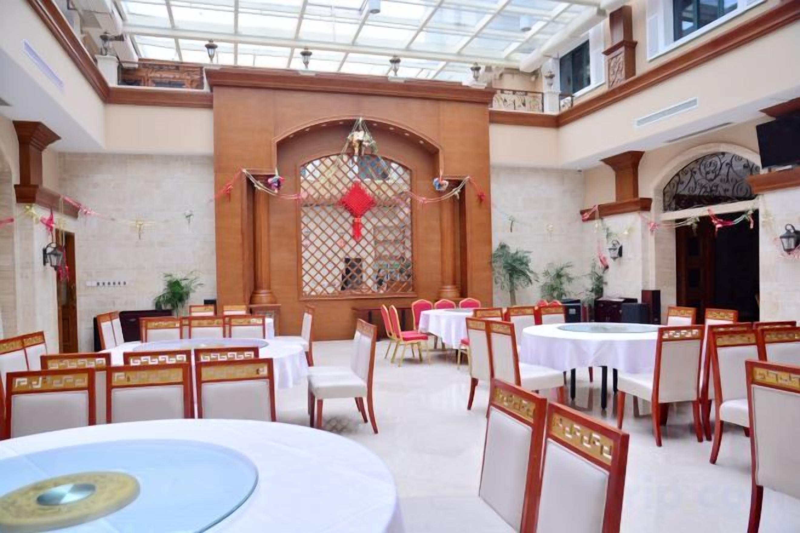 A banquet hall with round tables and chairs, a high ceiling with a skylight, and a decorative window with a red ornament.