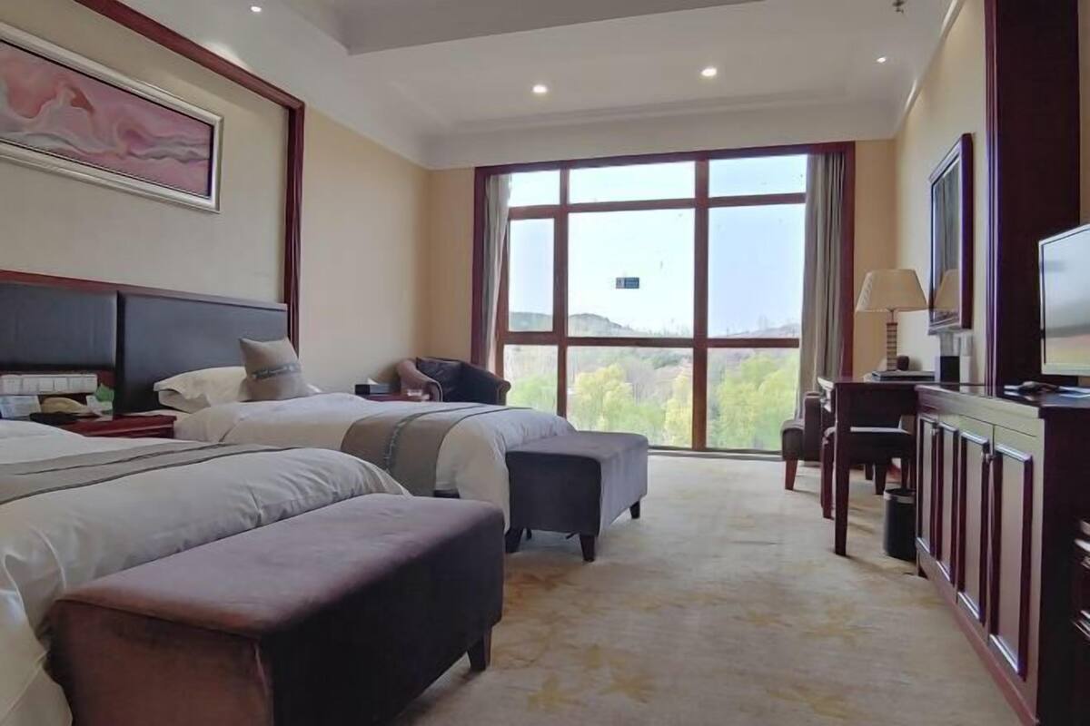 Ein Hotelzimmer mit zwei Betten, einem Schreibtisch, einem Fernseher und einem großen Fenster mit Blick ins Freie.