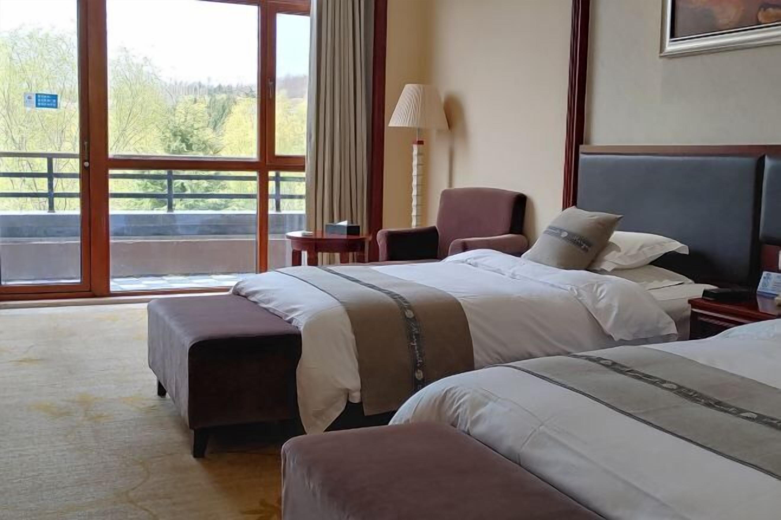 A hotel room with two beds, a chair, a lamp, and a view of the outdoors.
