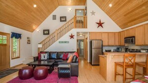 L’intérieur de la cabane comprend une chambre à coucher située en mezzanine, un salon avec un canapé et une table basse, une cuisine avec des meubles en bois, et un coin repas avec une table et des chaises.