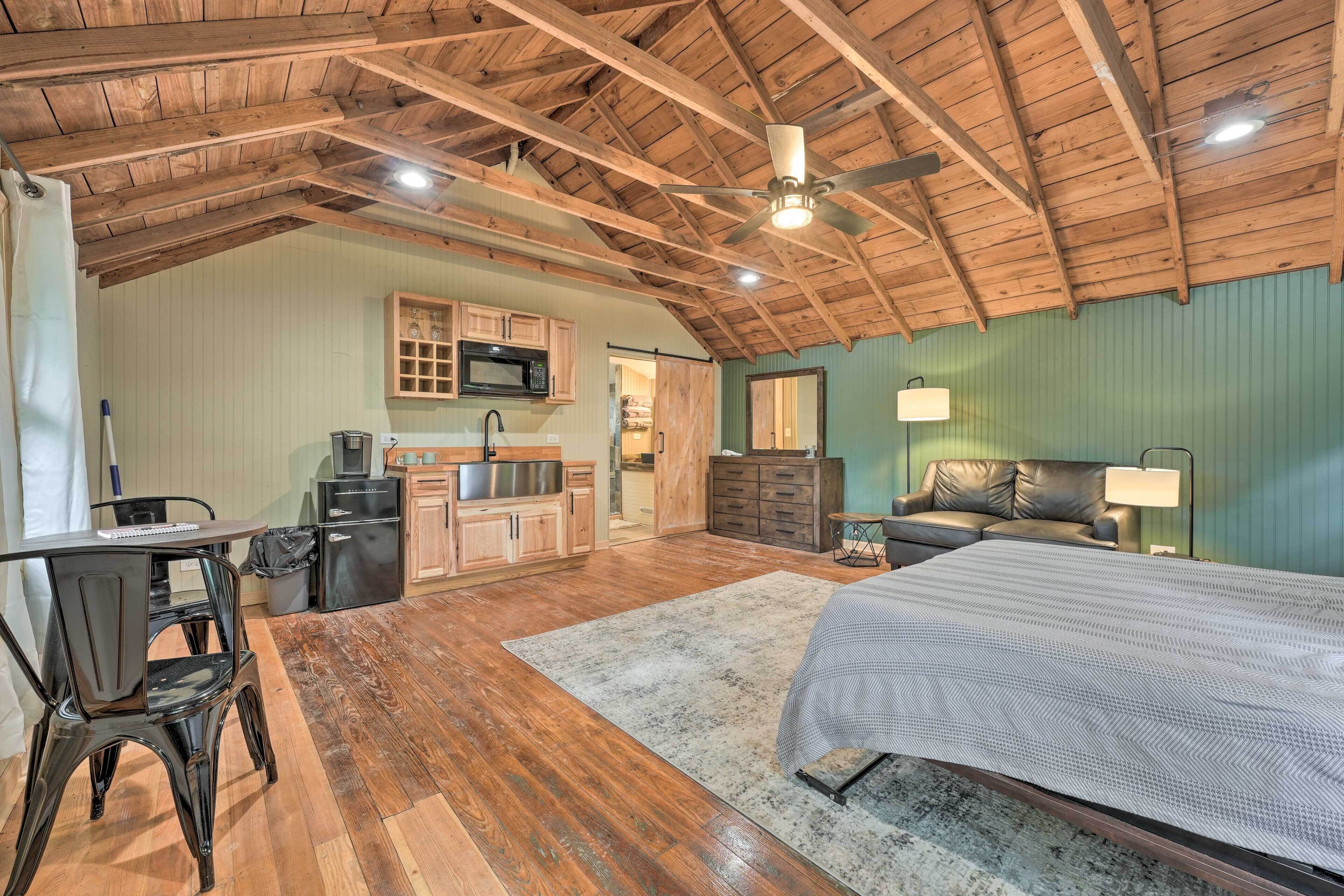A spacious room with a wooden ceiling, a bed, a desk with a chair, a kitchenette, and a bathroom.