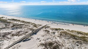 Vue aérienne d’une plage avec une promenade en bois menant au rivage, une eau d’un bleu limpide et des parasols de plage.