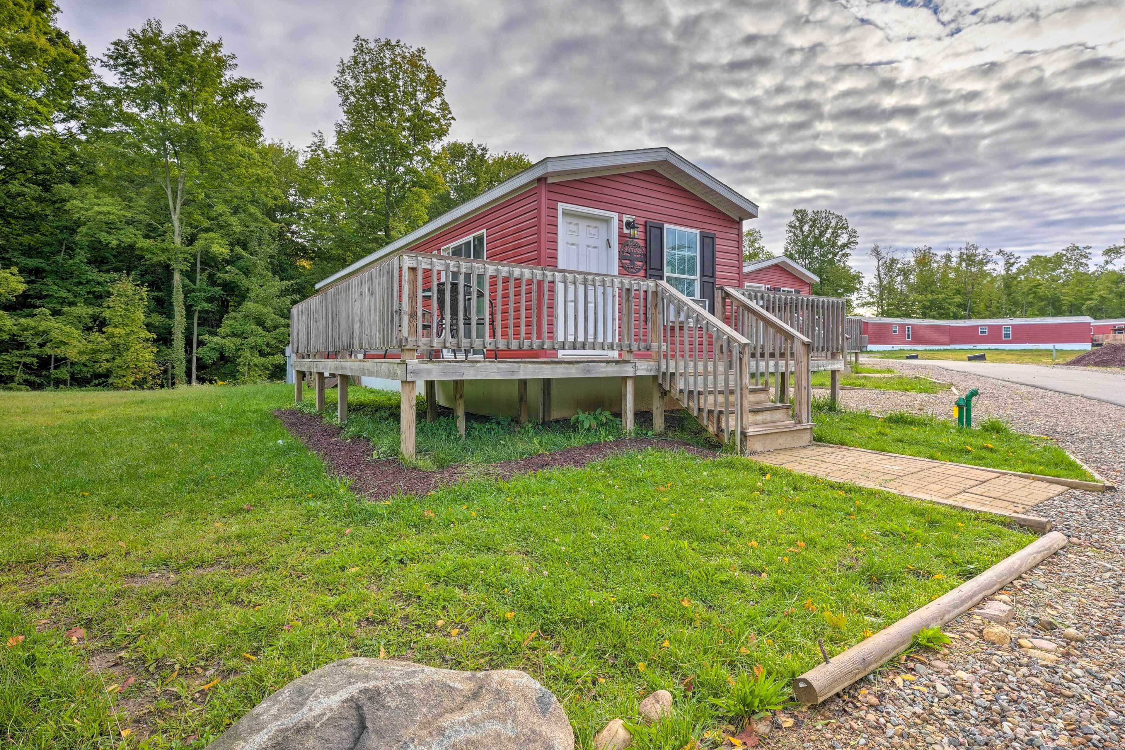 A red cabin with a wooden deck and stairs, surrounded by greenery and a cloudy sky.