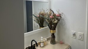 Kylpyhuoneessa on peili, pesuallas, nestesaippuapumppu, kuivien kukkien koristama maljakko ja peilin yläpuolella oleva valaisin.