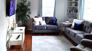 Een woonkamer met een grijze bank, een witte bijzettafel, een witte tv-meubel en een plafondventilator.