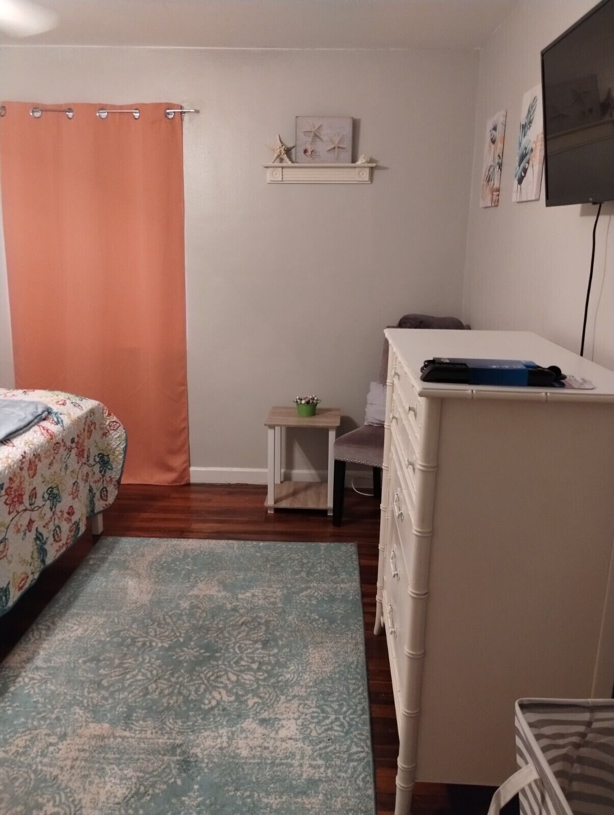 Une chambre à coucher comprenant un lit, une commode, une petite table avec une plante, une étagère avec des objets décoratifs et un téléviseur fixé au mur.