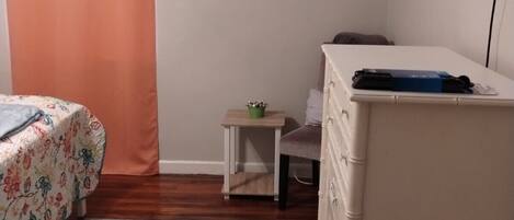 Une chambre à coucher comprenant un lit, une commode, une petite table avec une plante, une étagère avec des objets décoratifs et un téléviseur fixé au mur.