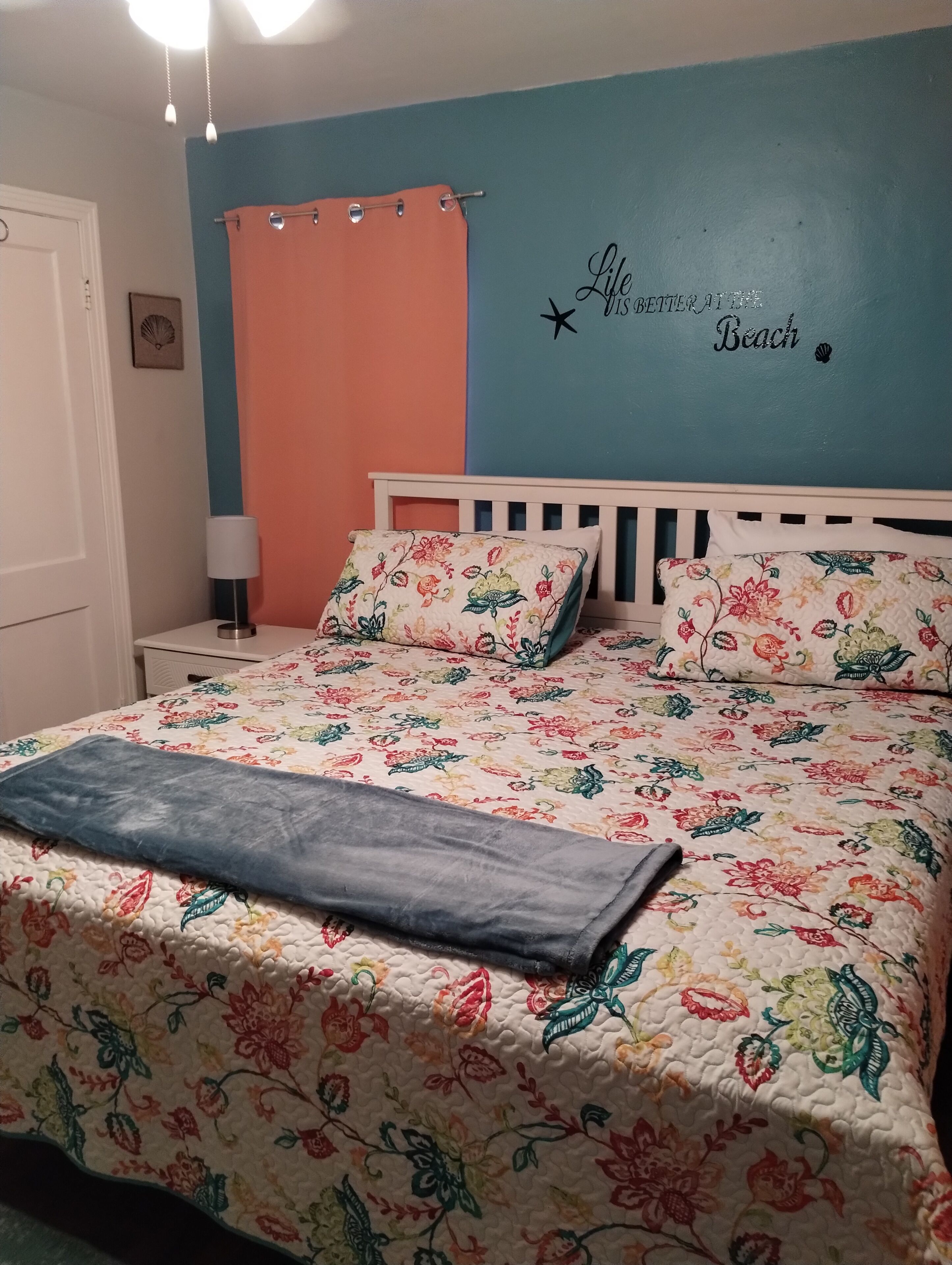 A bedroom with a floral bedspread, a white headboard, and a wall with a decorative phrase.