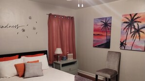 Een keurig ingerichte slaapkamer met een bed, nachtkastje, stoel, en twee kunstwerken die zonsondergangen en palmbomen voorstellen.