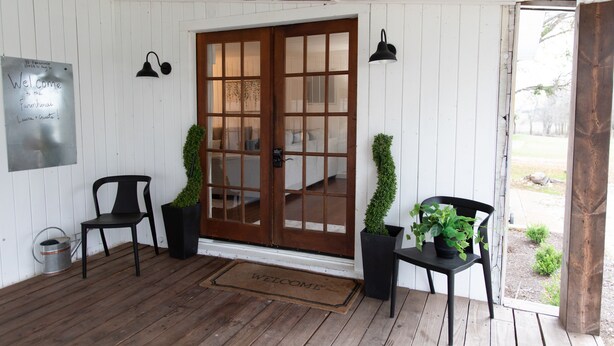 Een veranda met houten vloer, een welkomstmat, twee stoelen, een deur met ruitpanelen en een bord aan de muur.