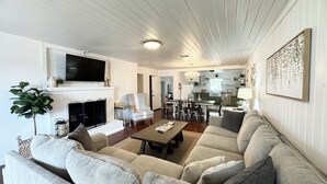 Een ruime woonkamer met een open haard, een televisie erboven en een eethoek op de achtergrond.