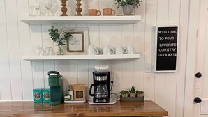 Een keuken met een houten aanrecht, twee witte barstoelen, een koffiezetapparaat en planken met bekers en een klok.