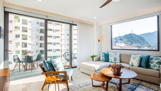 A modern living room with a sofa, chairs, a coffee table, and a large window offering a view of a cityscape and mountains. - BRAND NEW PENTHOUSE CONDO IN ROMANTIC ZONE - LISTEN TO THE WAVES (Puerto Vallarta)