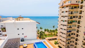A rooftop pool area with a view of the sea, adjacent buildings, and a palm tree-lined beach. - BRAND NEW PENTHOUSE CONDO IN ROMANTIC ZONE - LISTEN TO THE WAVES (Puerto Vallarta)