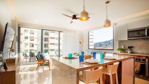 A modern kitchen with a large island, a dining area with chairs, and a view of the city through large windows. - BRAND NEW PENTHOUSE CONDO IN ROMANTIC ZONE - LISTEN TO THE WAVES (Puerto Vallarta)
