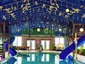 An indoor swimming pool with slides, a wooden deck, and a ceiling with hanging lights.
