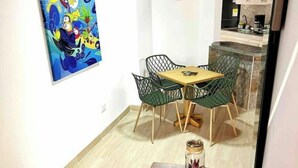 Uma área de refeições com uma mesa de madeira e cadeiras verdes, um quadro na parede e uma cozinha ao fundo.