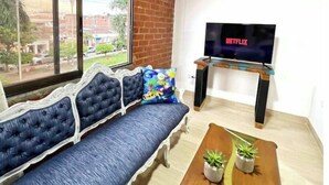 Uma sala de estar com um sofá, uma mesa de centro com plantas, uma TV em um suporte e uma janela grande com vista para prédios e árvores.