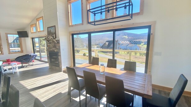 A spacious dining area with a large table and chairs, a stone fireplace, and large windows offering a view of a residential area. - Modern Cabin Facing Canaan Valley Ski Slopes (Davis)