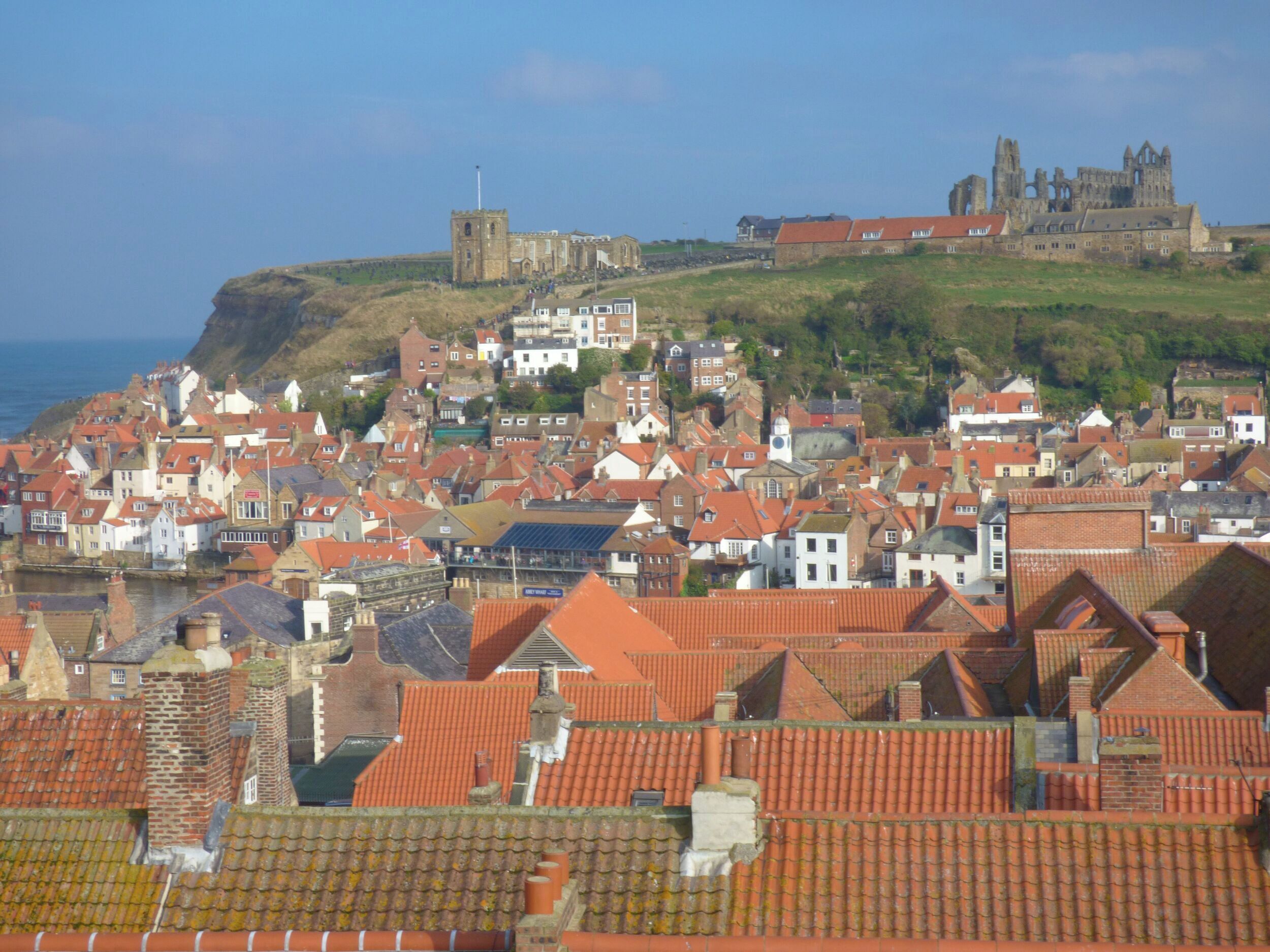 Une ville côtière aux toits de tuiles rouges, dotée de bâtiments historiques et surmontée d’un château imposant situé sur une colline.