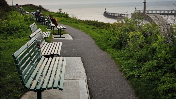 A coastal walkway with benches, a lighthouse, and a pier. - Whitby Haven (Whitby)