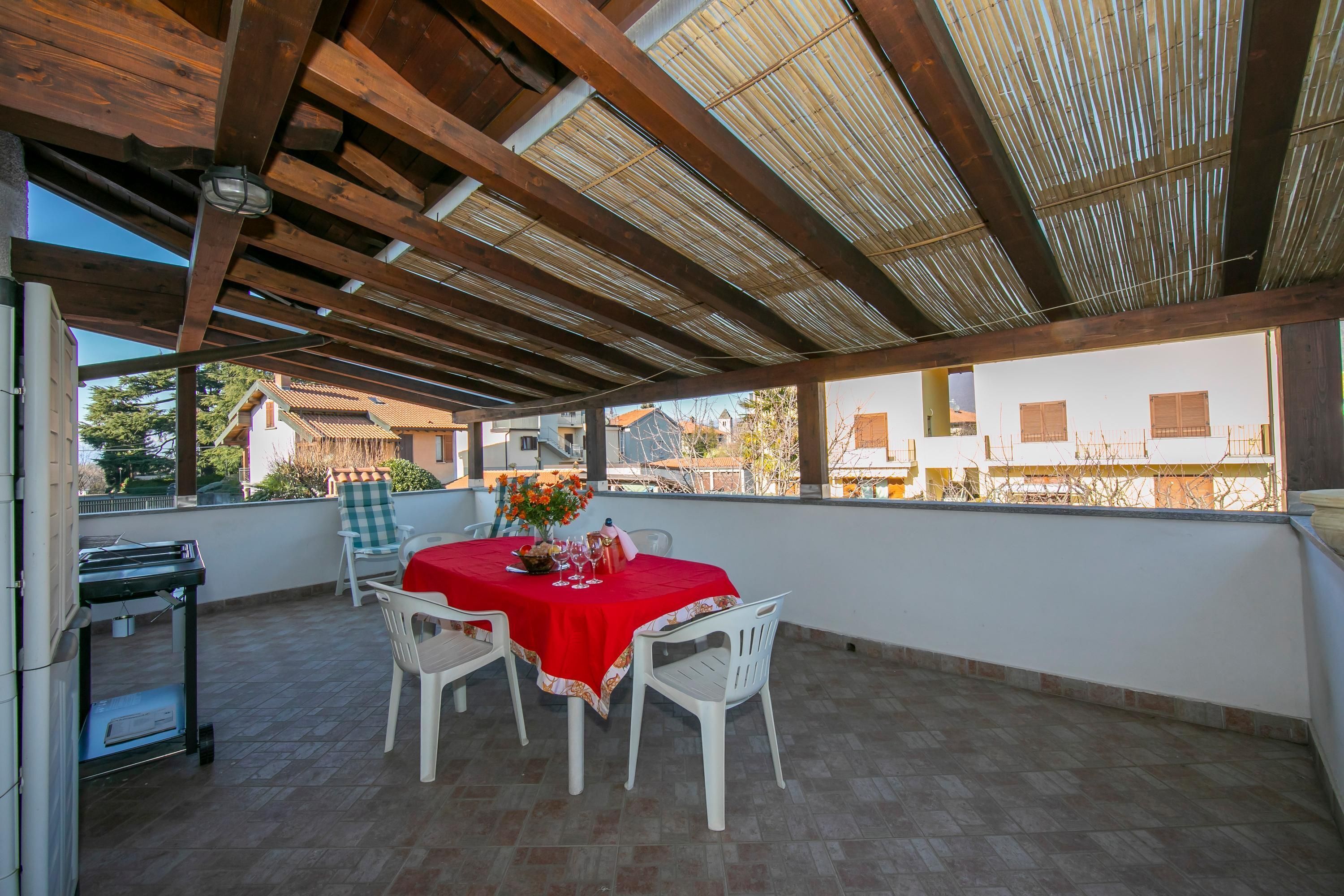 Une terrasse couverte avec une table dressée pour deux personnes, des chaises blanches et une nappe rouge.