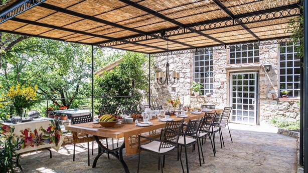 An outdoor patio with a wooden table set for dining, surrounded by chairs, under a thatched roof. - Casa Bosco a Stone Mansion in Chianti (Gaiole in Chianti)