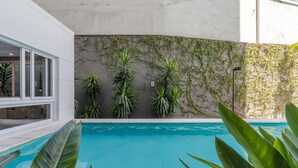Un área de piscina moderna con una piscina azul cristalina, rodeada de grandes plantas tropicales y un muro cubierto de enredaderas.
