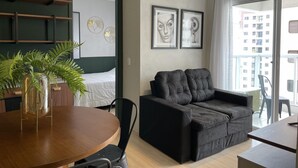 A modern living room with a black sofa, a wooden dining table, and a potted plant. There are framed artworks on the wall and a balcony with a view of buildings. - Hype055 in S o Paulo (São Paulo)