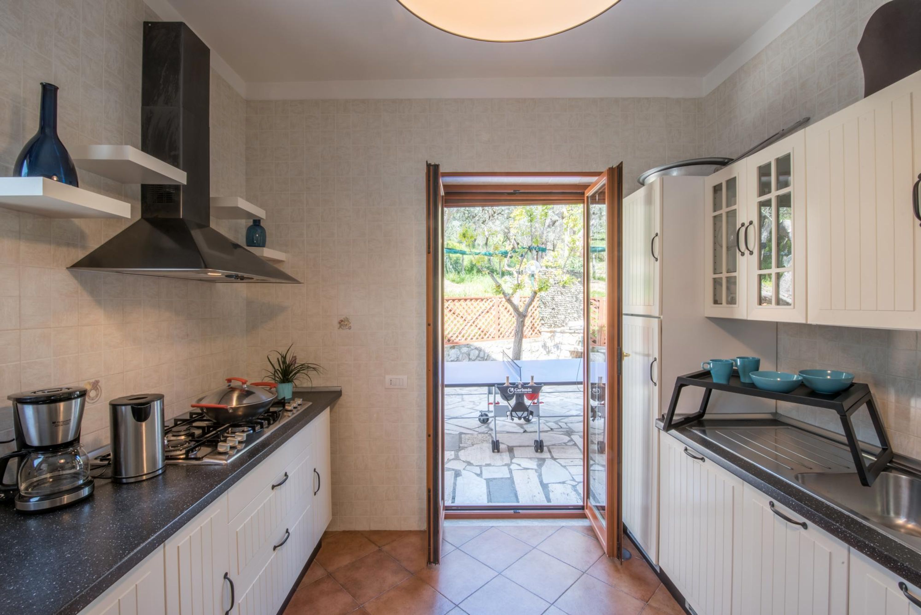 A modern kitchen with white cabinets, a stainless steel range, and a black range hood. There is a window leading to an outdoor area with a patio and a tree.