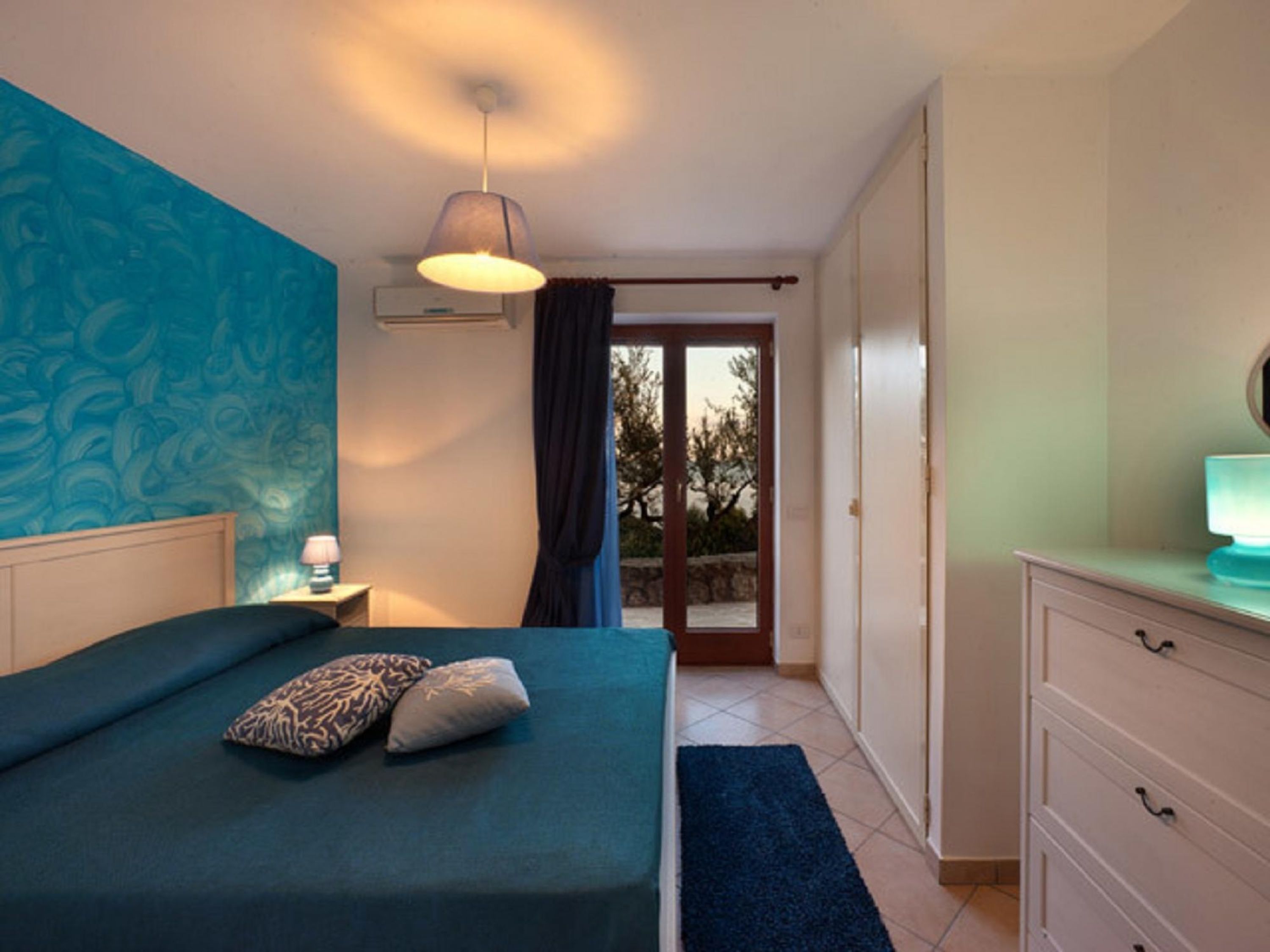 A bedroom with a bed, a nightstand, a dresser, and a view of the outdoors through a door.